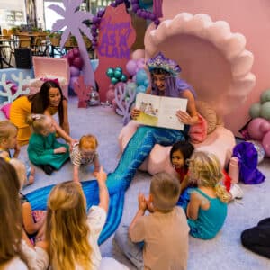 A mermaid performer reading a story book to children at a corporate event.