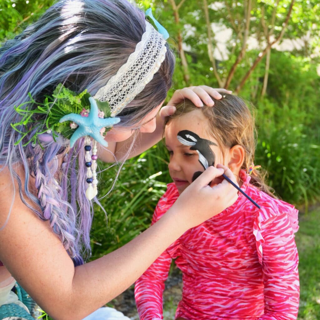 a mermaid performer applying face paint to a child's face