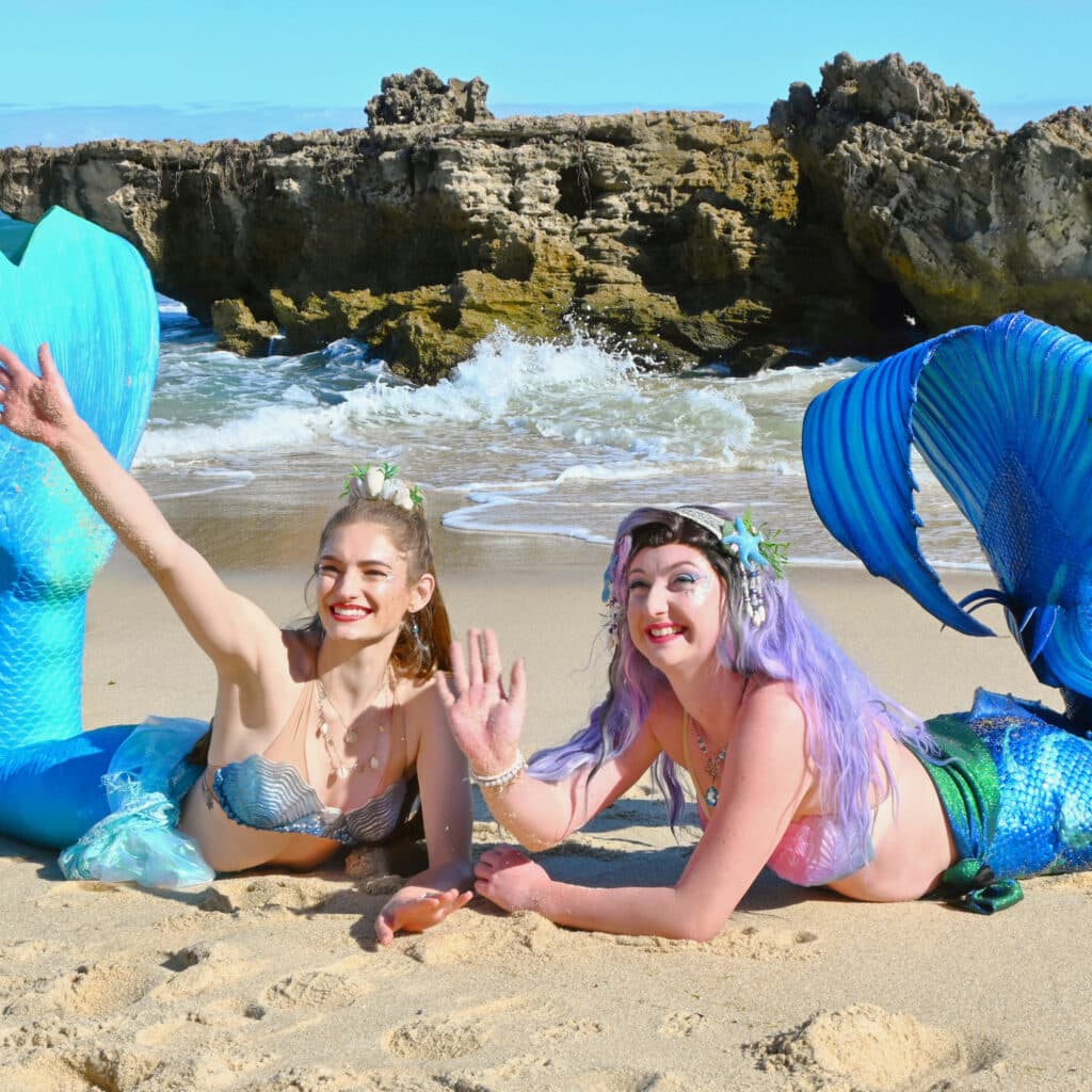Two mermaid performers in the sand at the beach, waving.