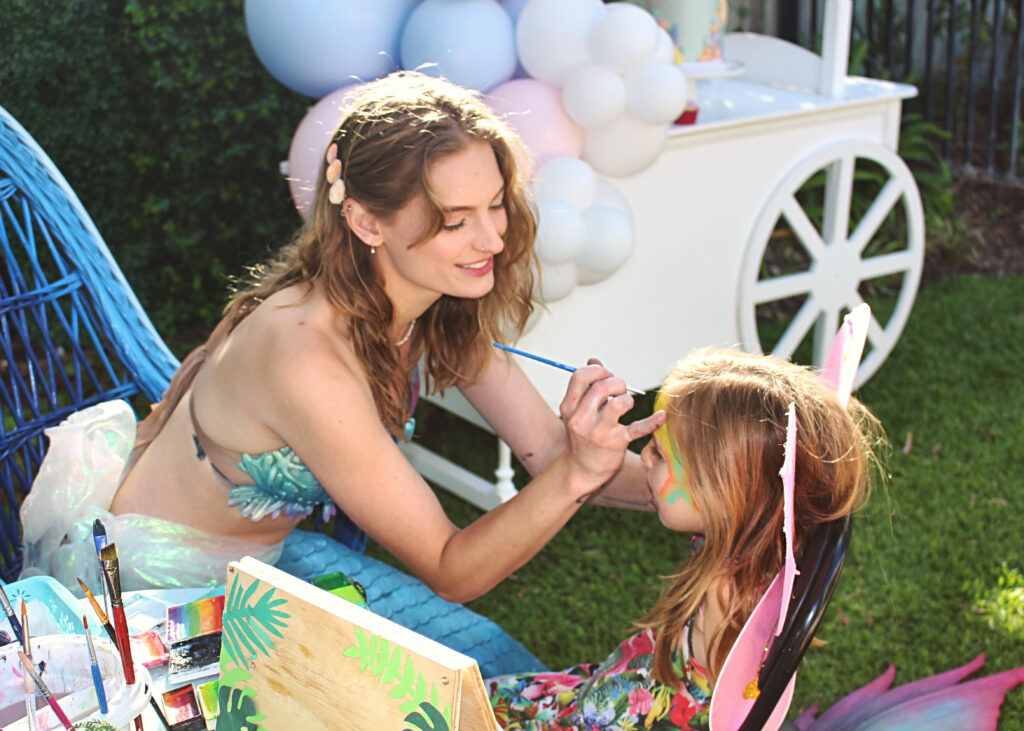 a mermaid performer applying face paint at a children's party